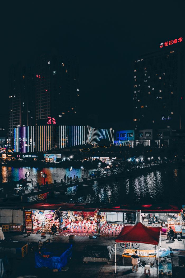 A bustling night market with vibrant lights by a modern cityscape reflected in water.