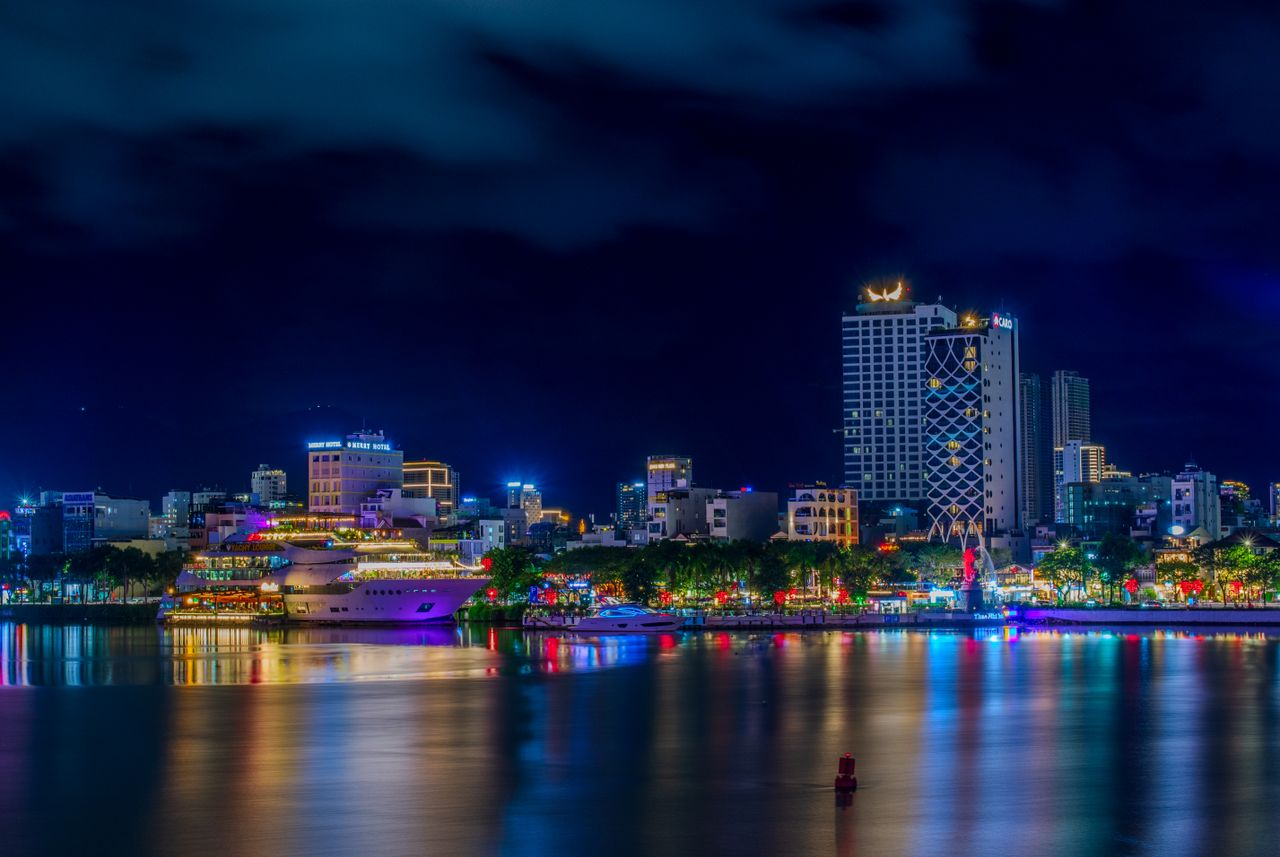 Vibrant night view of Da Nang skyline reflecting over the water with colorful lights.
